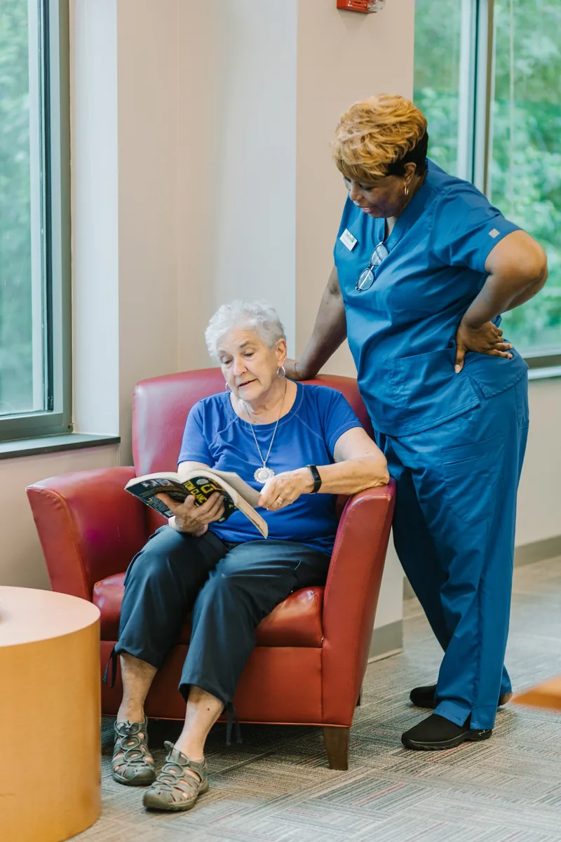 DayBreak caregiver standing beside an older adult reading in a chair